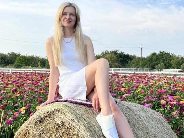 MelissaTyler with long blonde hair, white dress, and white socks, sitting on a hay bale in a colorful flower field, showing her bare legs and smiling.