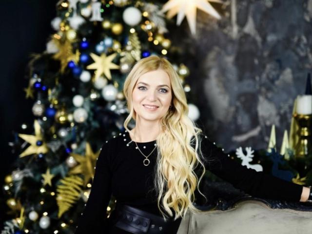 MelissaTyler with long blonde hair, black top, and necklace, smiling, standing next to a decorated Christmas tree with gold stars and blue ornaments, wearing a black belt.
