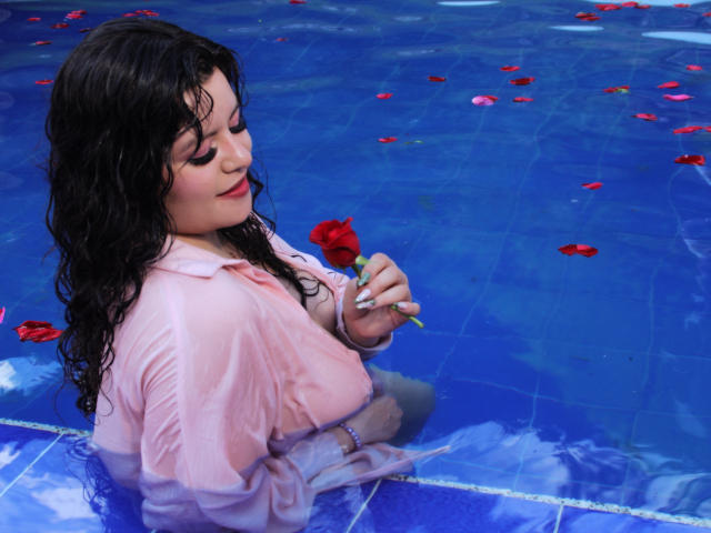 Sofinoa with wet black hair, holding a red rose, wearing a soaked pink shirt, sitting in a blue pool, surrounded by floating red rose petals.