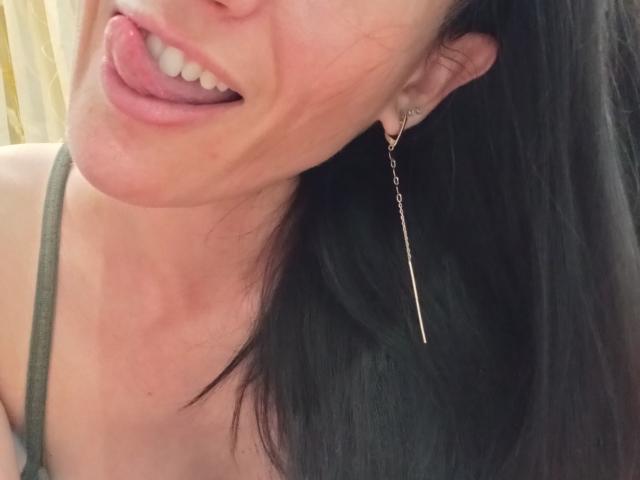 BellaJulls with tongue out, green tank top, black hair, and a long gold earring. Her tongue is touching her upper lip. Her tits are slightly visible.