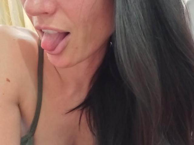 BellaJulls' face with tongue out, showing a bit of her upper chest and green strap. She has long dark hair, fair skin, and small dark moles on her shoulder.