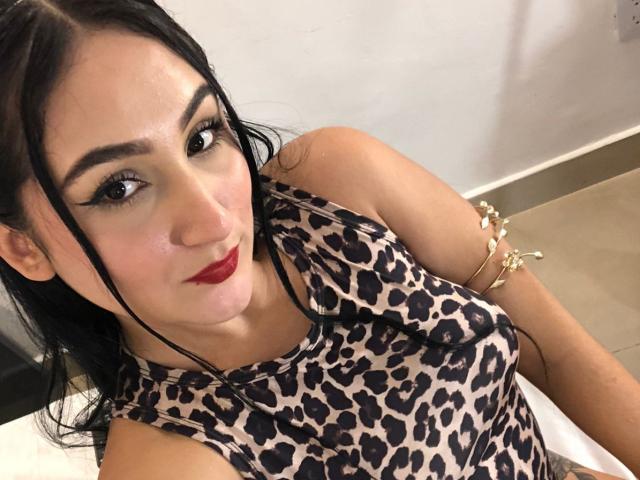HiilaryDolce with dark hair, red lipstick, and a leopard print top, showing her tits, wearing a gold arm bracelet, and looking at the camera with a slight smile.