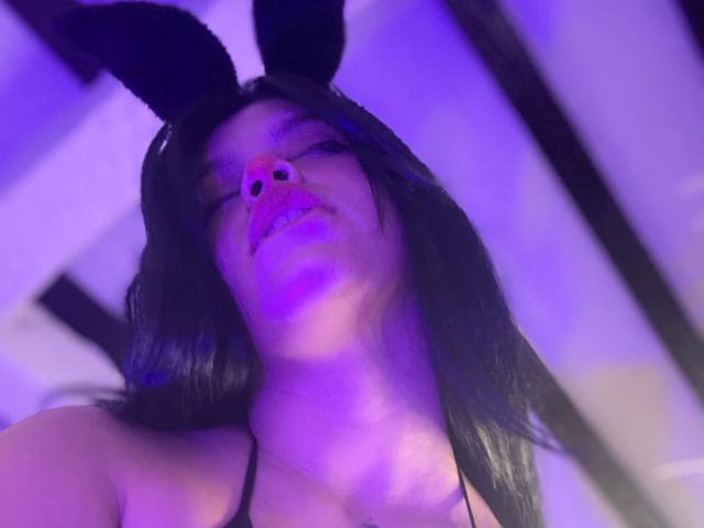 AryButty with dark hair, black bunny ears, and a nose clip, looking up with purple lighting. Boobs visible, wearing a black top. Low-angle shot.