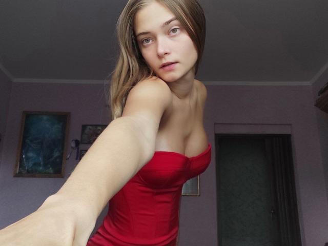 HappyEmily with long brown hair, green eyes, red strapless dress, small tits, and a slight smile, taking a selfie in a purple-walled room. Her right arm is extended.