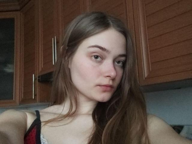HappyEmily has long brown hair, a slightly puffy nose, and a neutral expression. She's wearing a black and red spaghetti-strap top, showing her small tits.