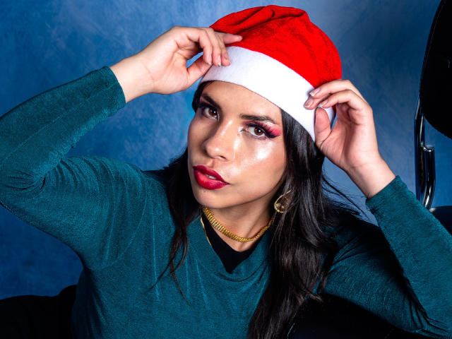 VioletaStone wearing a red Santa hat, green sweater, and gold chain, with pink eyeshadow, glossy red lips, and long dark hair, adjusting the hat with both hands.