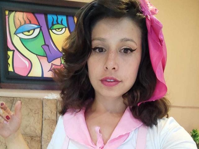 SarahWelinton has medium brown wavy hair, pink lipstick, and a pink flower in her hair. She's wearing a pink and white shirt, with eyeliner and red nails.