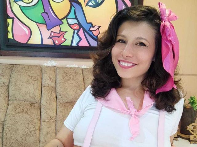 SarahWelinton with curly brown hair, pink bow, white shirt, pink scarf, smiling, showing teeth, cute, small tits, soft pink lipstick, seated, colorful painting in background.
