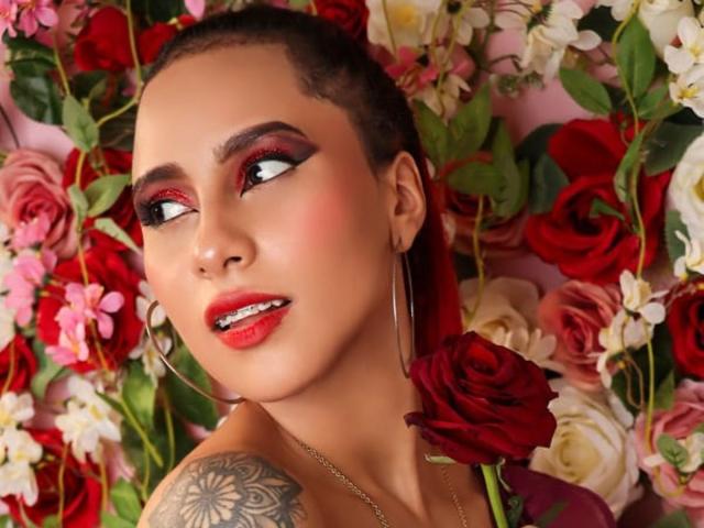 LeahMiler with red lipstick, bold eye makeup, and hoop earrings, holding a red rose, has a tattoo on her shoulder and a slight smile, surrounded by roses.