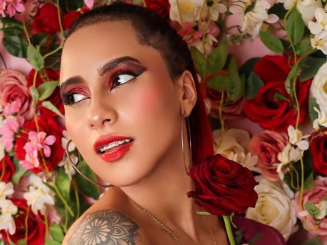LeahMiler has red lipstick, bold eyeliner, and a tattoo on her right shoulder, looking up with a flower in her hair and roses around her. She's wearing hoop earrings.