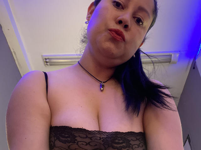 MarianaSmitt with black lace top, showing her tits, has medium-sized boobs, dark hair in a ponytail, wearing a black necklace with a purple stone, and red lipstick.