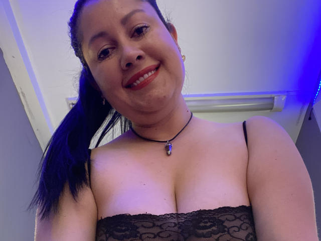 A photograph of MarianaSmitt with light skin and black hair in a ponytail. She has a broad smile, red lipstick, and wears a black lace strapless top that reveals cleavage. She has small earrings and a black necklace with a purple pendant. The image is taken from a low angle, emphasizing her upper body and face. The background is mostly out of focus, with a blue light on the right. MarianaSmitt's facial features are clear, and her expression is friendly.