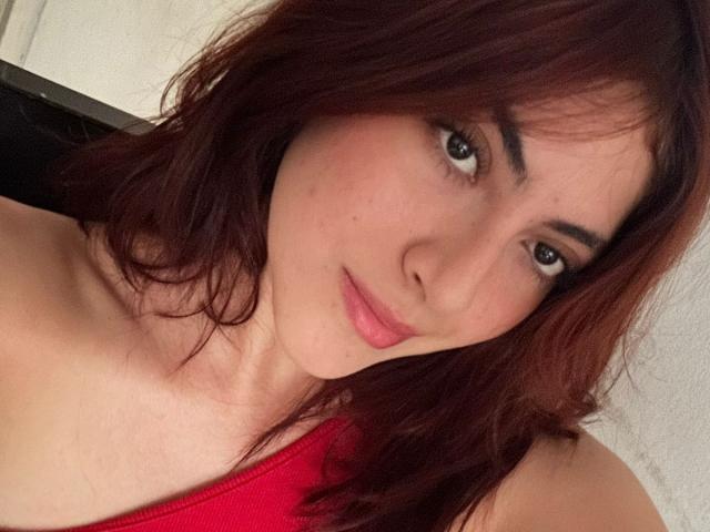 SpencerLe has reddish-brown hair, light skin, and a subtle smile with pink lips. She's wearing a red top, showing her shoulders and upper chest. She has small tits.