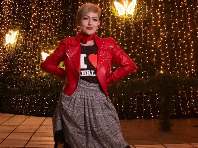 MaferCougar stands in a nighttime scene with a dark, string-lit backdrop. She wears a red leather jacket with silver studs over a black T-shirt with "I ❤️ Berlin" text. Her gray, patterned skirt flows down to her ankles. She has blonde hair, red lipstick, and a confident, slightly smug expression. Three glowing butterfly lights are in the background. The image has a casual, street-style vibe.