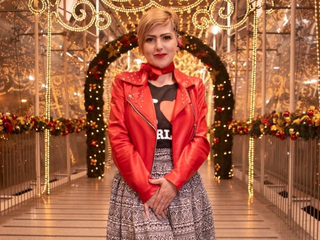 MaferCougar stands in a festive, illuminated corridor. She has short blonde hair, red lipstick, and a confident expression. She wears a red leather jacket with black text "BLK" on a black top, and a gray patterned skirt. Her hands are clasped in front. The background features ornate, gold-patterned walls, Christmas decorations, and string lights. The image has a warm, festive color palette. MaferCougar's stylish, bold outfit contrasts with the elegant, festive background.