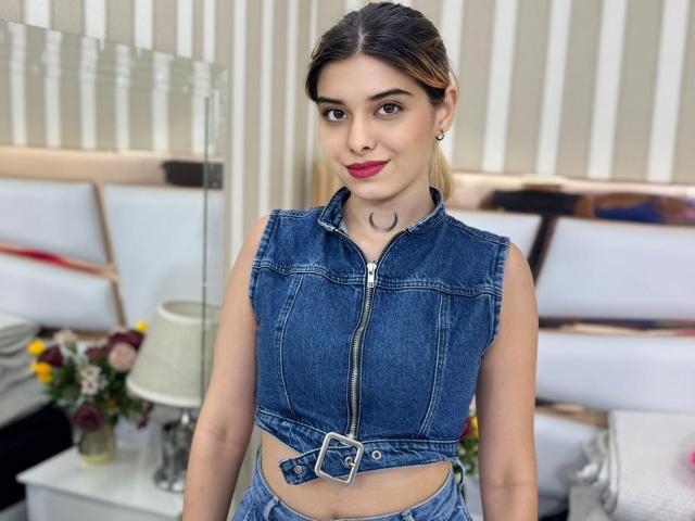 Lanna Hamilton, à la peau claire, aux cheveux bruns foncés, avec du rouge à lèvres, porte un top en jean bleu sans manches avec une fermeture éclair sur le devant et une ceinture, a de petites seins, et se tient dans une chambre..