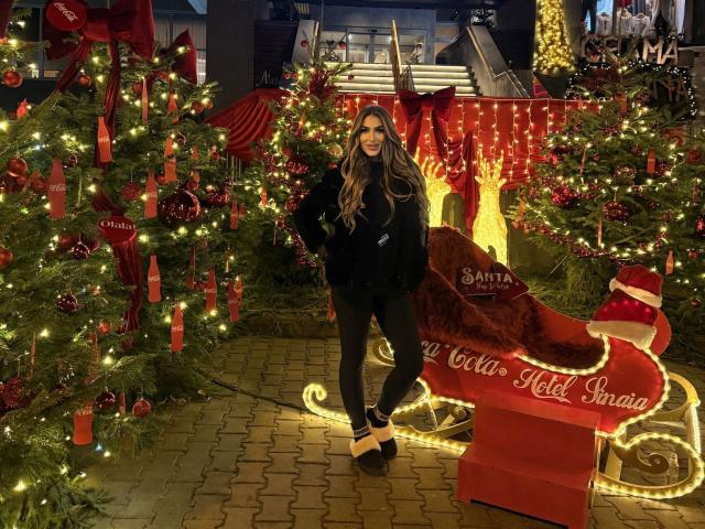 NicolSNaya stands in a Christmas display with glowing trees, red ornaments, and a red sleigh, wearing a black fur coat, black pants, and white heels, with long brown hair.