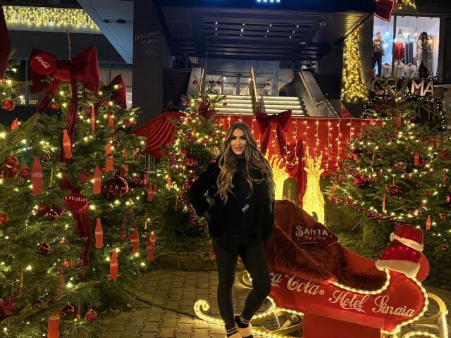 NicolSNaya stands in front of Christmas trees, glowing lights, and a red sleigh, wearing a black jacket, black pants, and sneakers, with long brown hair and a smile.
