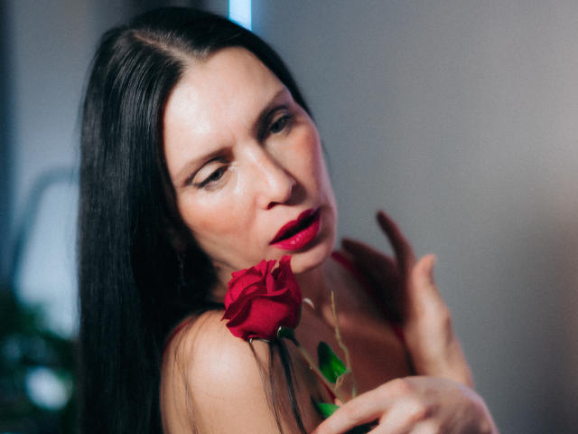 EvanessaJohn with long black hair, red lipstick, and a red rose in her hand, looking seductively at the camera, her tits slightly visible in a red top.