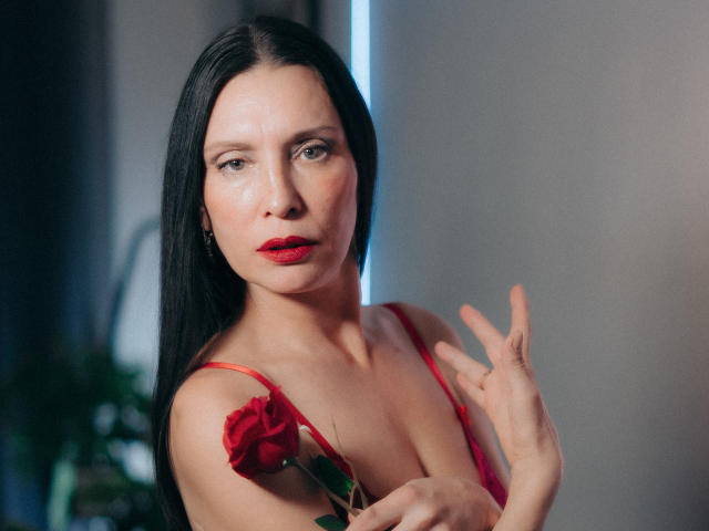 EvanessaJohn with long black hair, red lipstick, holding a red rose, wearing a red spaghetti strap top, raising her right hand, looking at the camera with a serious expression.
