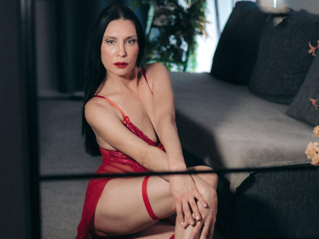 EvanessaJohn with long black hair, red lingerie, and red garter, sitting on a gray couch, showing her boobs, with red lips and a serious expression.