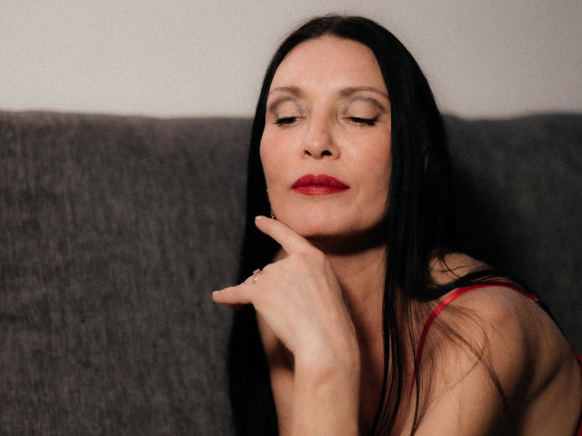 EvanessaJohn with long black hair, red lipstick, and closed eyes, touching her chin with a finger, wearing a red top, sitting on a gray couch.