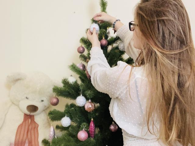 KarKutie with long brown hair, wearing glasses and a white polka-dot shirt, decorating a Christmas tree with pink and white ornaments, next to a large white teddy bear.