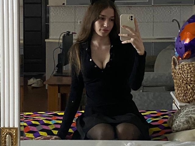 KaiaLein met lang bruin haar, een zwarte blousejurk, waarbij haar borsten zichtbaar zijn, zittend op een kleurrijk bed, een spiegel-selfie makend met een iPhone, zwarte kousen aan, een rieten mandje aan de rechterkant..