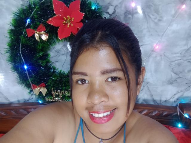 MariahSquirting69 with dark hair, brown eyes, and pink lipstick, smiling, topless, wearing a blue halter, Christmas wreath with poinsettias and lights behind her.