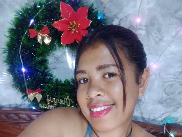 MariahSquirting69 with dark brown hair, medium skin, red poinsettia flower, green Christmas wreath, and blue lights, smiling with pink lipstick, wearing a blue halter top, in a selfie.