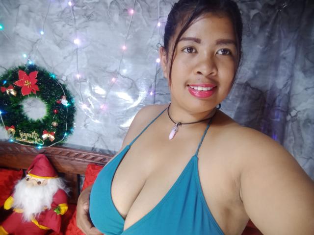 MariahSquirting69 with tan skin, dark hair in a bun, wearing a teal halter top showing big tits, pink lipstick, black choker, smiling, standing with a Santa doll.