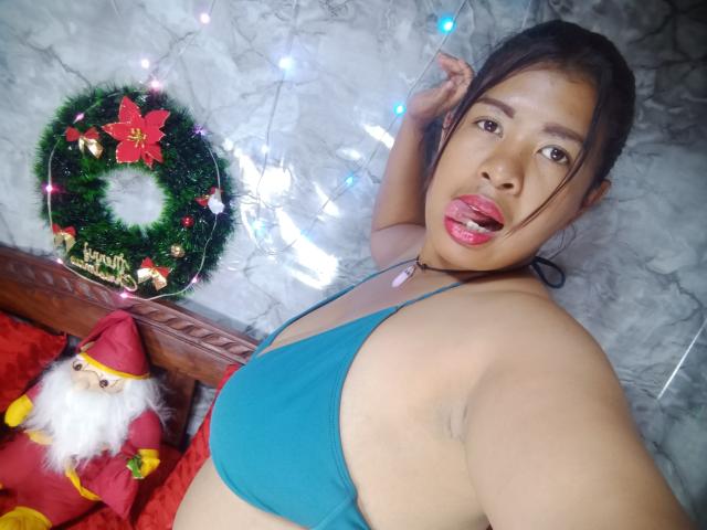 MariahSquirting69, Latina with tan skin, black hair in ponytail, blue bikini top, tongue out, looking at camera, Christmas wreath, Santa doll, white string lights in background.