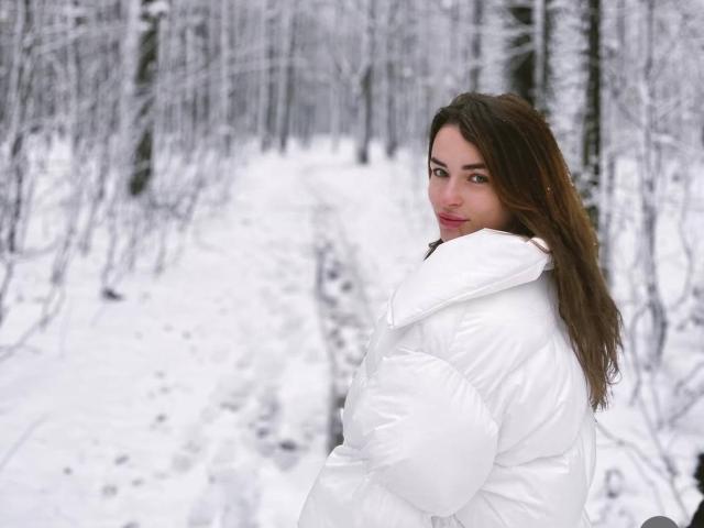MandiBrooks with long brown hair, wearing a puffy white jacket, looking over her shoulder with a slight smile, standing in a snowy forest. Her tits are partially visible.