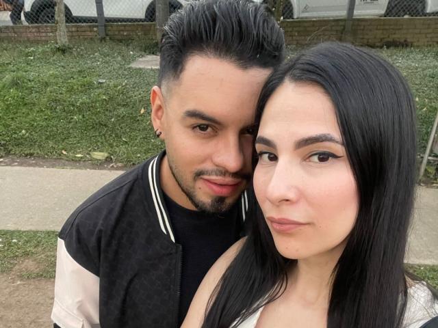 AriannaAndDave with dark hair, makeup, and a white top, has a serious expression. Dave, with a beard and black jacket, stands behind her, leaning in.