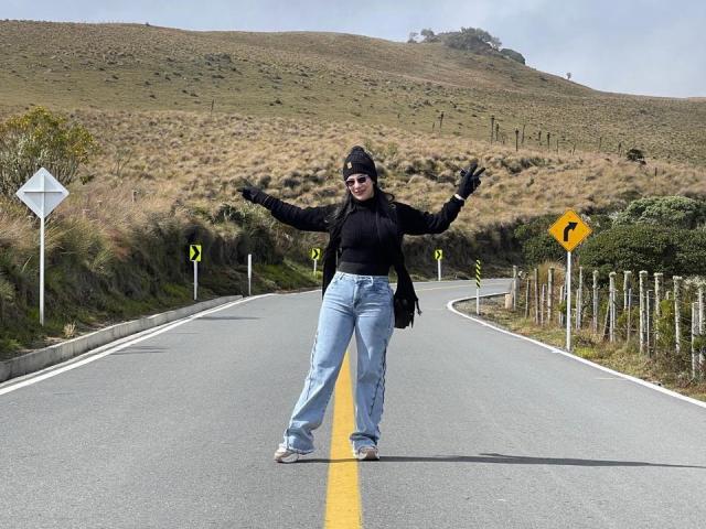 AriannaAndDave stands on a road with arms out, wearing a black top, jeans, and beanie, smiling with arms spread, in a sunny, hilly landscape.