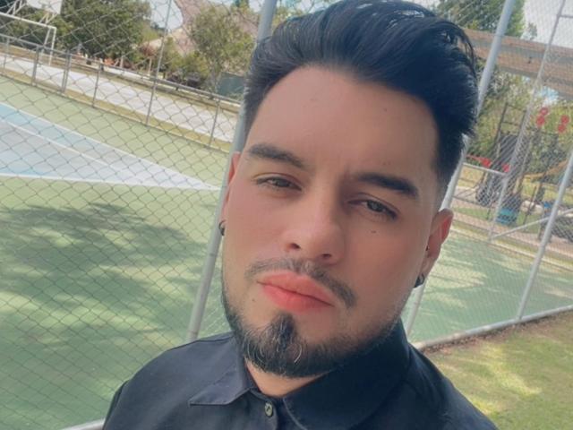 AriannaAndDave, with dark hair, a beard, and black ear gauges, wearing a black shirt, stands in front of a tennis court, showing a serious expression.