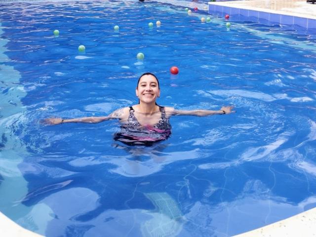 AliRousse with tattoos, blue swimming pool, arms out, smiling, wearing a pink and black swim top, floating with colorful buoys in the background, water reflecting light.