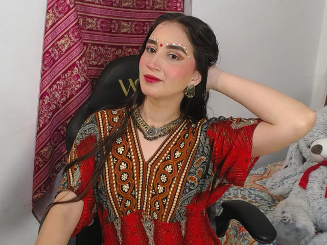 JalimaMona with long dark hair, red and brown patterned dress, gold jewelry, and a facial decoration, smiling, hand in hair, sitting in a black chair, fluffy gray toy.