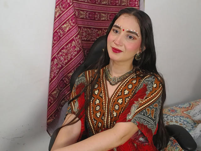 JalimaMona with long dark hair, red and brown patterned dress, white face dots, red lipstick, gold necklace, and earrings, sitting in a black chair, looking slightly to the right.