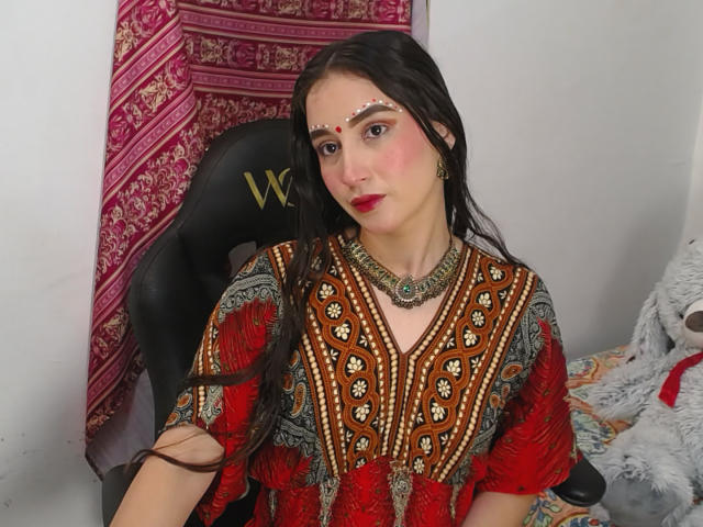 JalimaMona with long black hair, red lipstick, and a traditional, ornately-patterned red and brown dress, wearing a jeweled necklace and facial jewelry, sitting in a black chair.