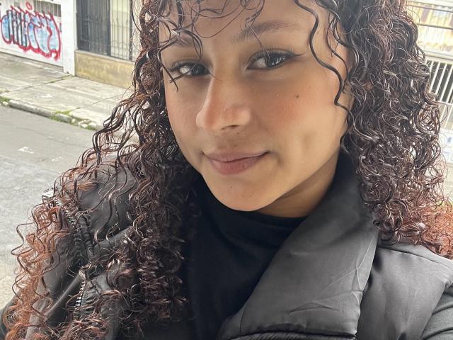 SofiaPassionne with curly brown hair, wearing a black turtleneck and gray puffy jacket, has a cute smile and subtle makeup, her brown eyes looking at the camera.