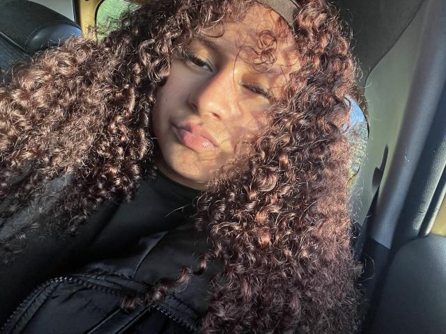 SofiaPassionne with curly brown hair, light brown skin, wearing a black puffy vest, winking with sunlight on her face, sitting in a car, looking cute and relaxed.