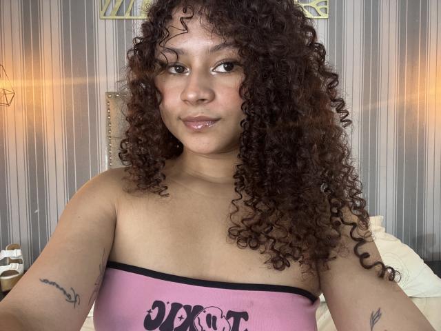 SofiaPassionne with curly brown hair, light brown skin, and small tattoos on her arms, wearing a pink strapless top, smiling slightly. Boobs visible, bed in background.