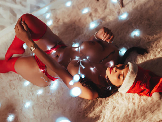 YelineMourine with big boobs, red panties, and red thigh-highs, lying on a white fluffy rug, wearing a Santa hat, with Christmas lights on her, and a tattoo on her chest.