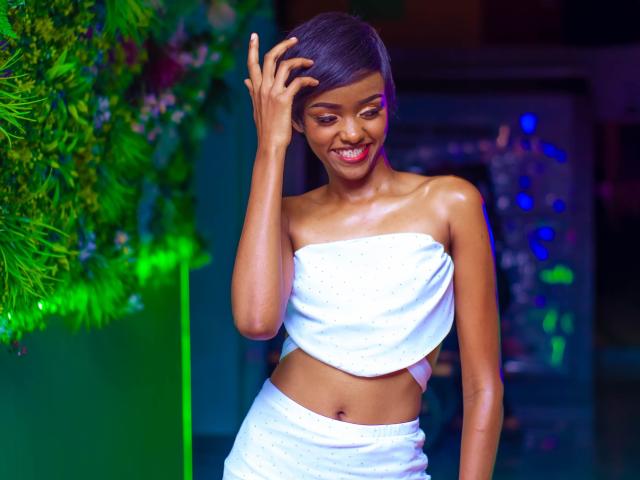 Sexyshuxy with dark skin, short black hair, smiling, wearing a white strapless top and matching skirt, touching her head, showing a slim, toned, shiny midriff.