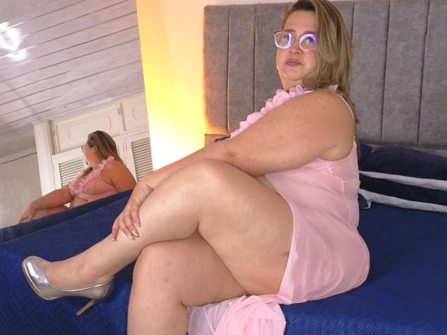CurvyRosse with glasses, pink sheer dress, and silver heels, sitting on blue bed, legs crossed, reflection in mirror, showing her curvy ass and plump thighs, blonde hair.