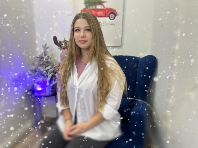 SweetDaylight with long blonde hair, white button-up shirt, and pink tee, sitting on blue chair, snowflakes in foreground, blue Christmas tree light on left, relaxed expression.