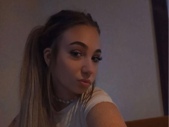 LilyLiebe-hot with puffy brown ponytail, thick eyebrows, long eyelashes, choker, and white shirt, looking over shoulder with a slight smirk. Dimly lit, soft brown skin.