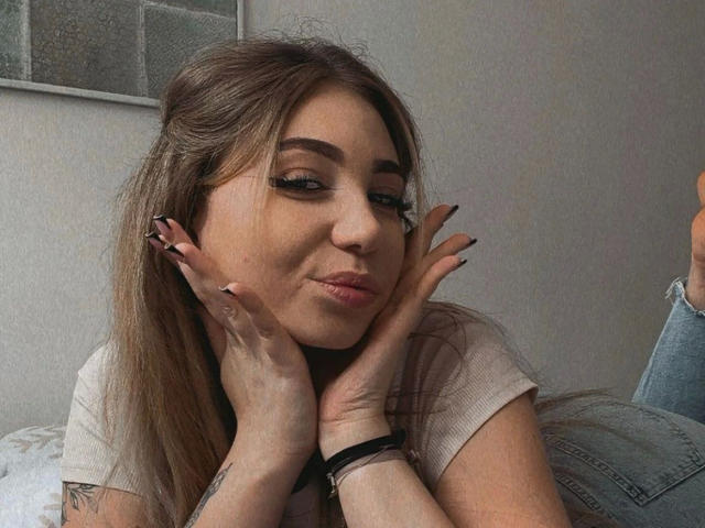 LilyLiebe-hot with long brown hair, full lips, and black eyeliner, pouting while resting her hands on her cheeks, wearing a beige top, showing her nails and wrist.