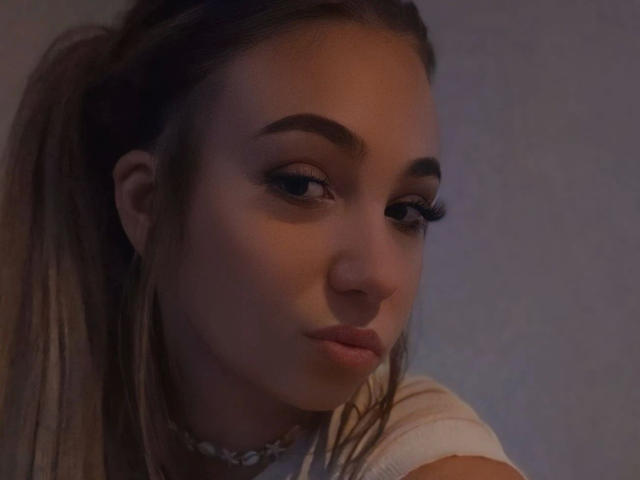 LilyLiebe-hot with pouty lips, dark eyebrows, long eyelashes, and brown hair in a ponytail, wearing a shell choker, looking slightly down. Soft, dim lighting.